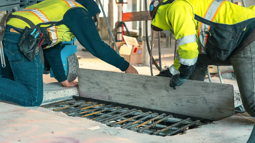 Two construction workers place a wooden board over exposed rebar during a slab opening in a concrete structure.