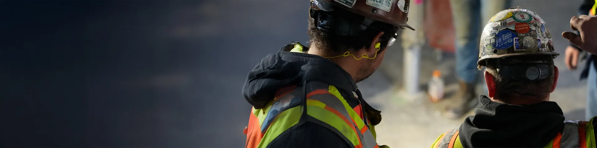 Two construction workers in safety vests and hard hats talk at a job site, one pointing toward an unseen area.