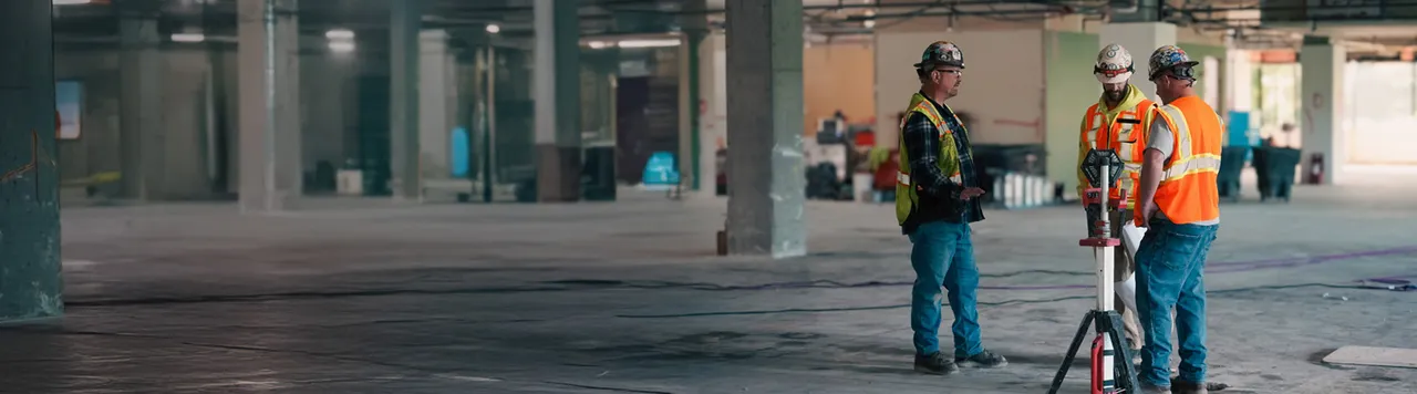 Three construction workers in safety gear discuss a project inside an unfinished commercial building.