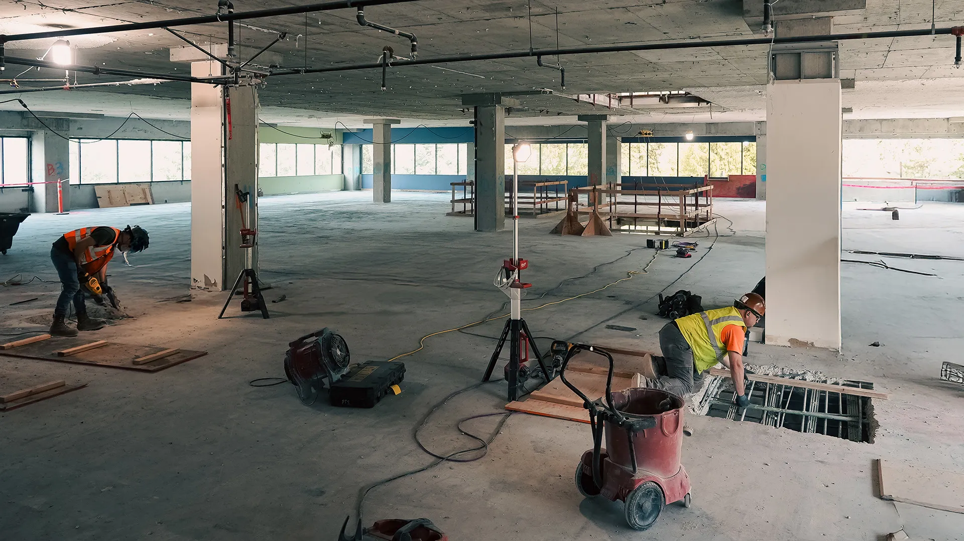 Two construction workers perform slab modifications in a large open building interior with exposed rebar and construction tools.