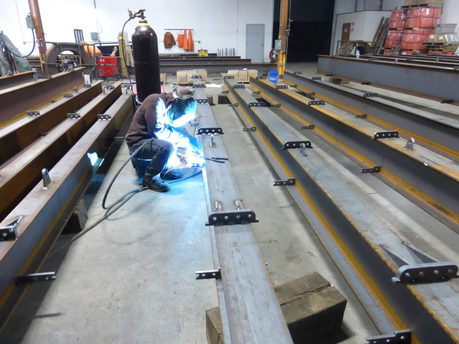 Worker welding connection plates onto steel I-beams inside a fabrication shop for Fortis Pile installation.