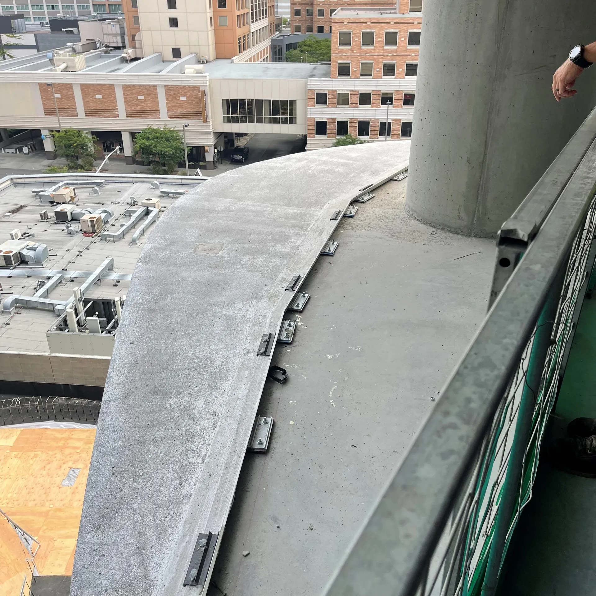 Curved concrete balcony on high-rise with exposed anchors and railing overlooking rooftop units.