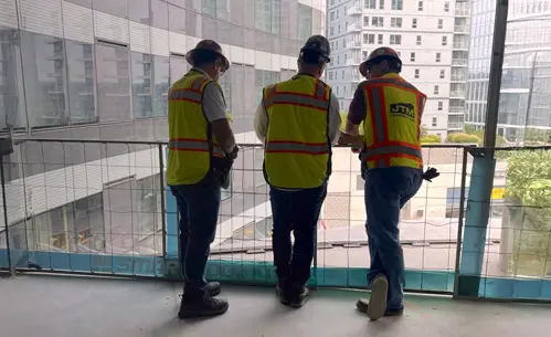 Three construction workers in safety vests stand on an upper floor of a high-rise, reviewing project progress onsite.